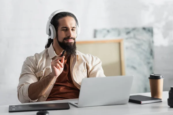 Designer latino em fones de ouvido segurando estilete perto de tablet gráfico borrado e laptop — Fotografia de Stock