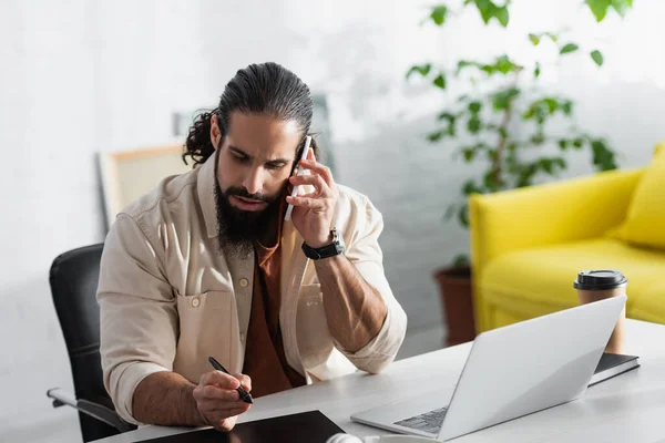 Freelance ispanico che tiene lo stilo vicino al tablet grafico mentre parla sul telefono cellulare — Foto stock