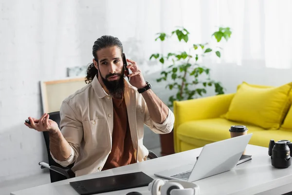 Retoucher hispano gesticulando mientras habla en smartphone cerca de laptop, tablet gráfica y cámara digital - foto de stock