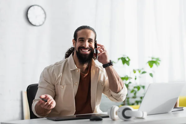 Diseñador sonriente hablando en el teléfono móvil cerca de la computadora portátil borrosa y auriculares - foto de stock