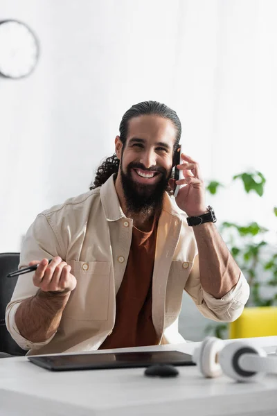 Designer feliz segurando stylus perto de tablet gráfico enquanto conversa no smartphone em casa — Fotografia de Stock