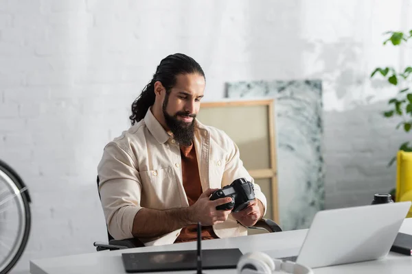 Spanischer Fotograf hält Digitalkamera in der Nähe von verschwommenem Laptop und Grafik-Tablet zu Hause — Stockfoto