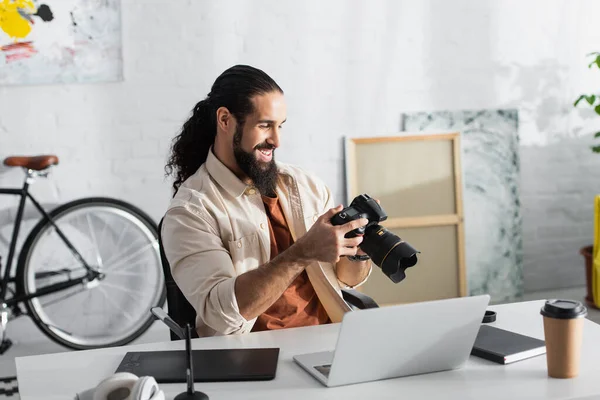 Felice fotografo ispanico in possesso di una moderna fotocamera digitale vicino al computer portatile e tablet grafico a casa — Foto stock