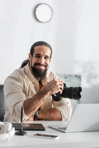 Fotógrafo feliz con cámara digital sentado cerca borrosa portátil y tableta gráfica en casa - foto de stock