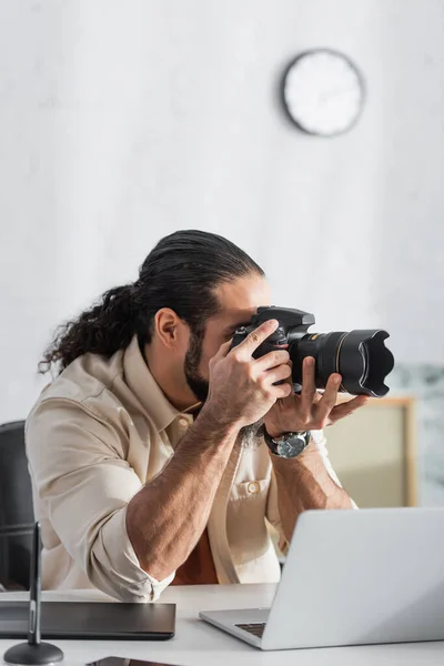 Hombre hispano tomando fotos en cámara digital cerca de un ordenador portátil borroso en casa - foto de stock