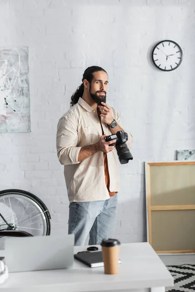 Вдумчивый латиноамериканец трогает бороду и смотрит в камеру, стоя дома с цифровой камерой — стоковое фото