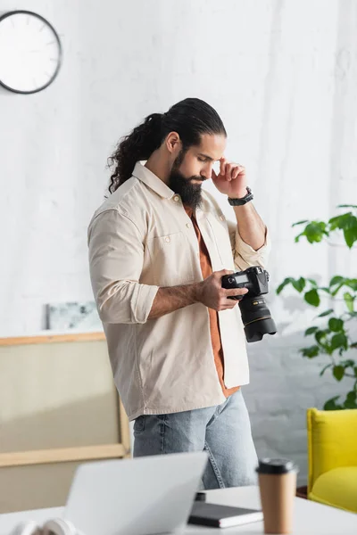 Pensativo fotógrafo hispano agarrado de la mano cerca de la cabeza mientras mira la cámara digital en casa - foto de stock