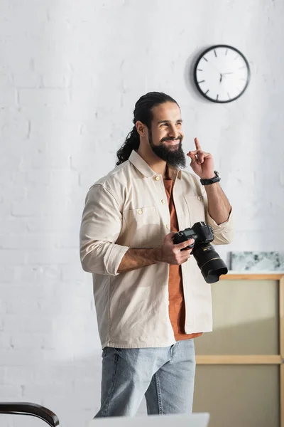 Веселый латиноамериканец с цифровой камерой, показывающий знак идеи, стоя дома — стоковое фото