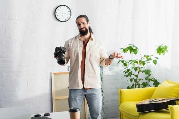 Confundido hombre hispano con cámara digital mostrando gesto encogiéndose de hombros mientras está de pie cerca de la guitarra y el sofá amarillo - foto de stock