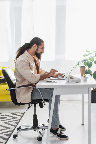 Side view of bearded latin freelancer typing on laptop while working at home — Stock Photo