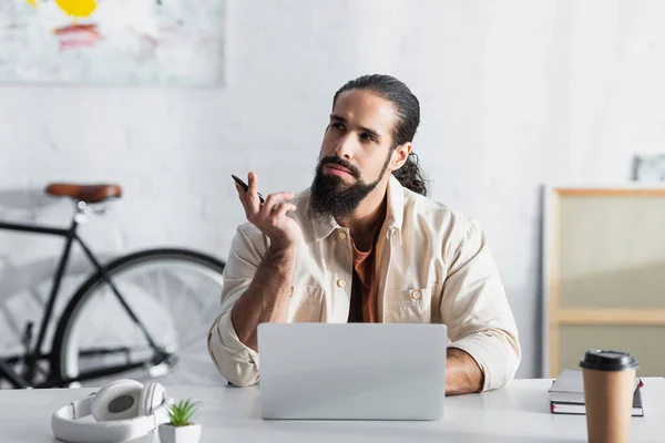 Pensativo independiente latino mirando hacia otro lado cerca de la computadora portátil en el lugar de trabajo - foto de stock