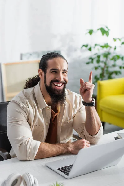 Alegre freelancer hispano apuntando con el dedo mientras trabaja cerca del portátil en casa - foto de stock