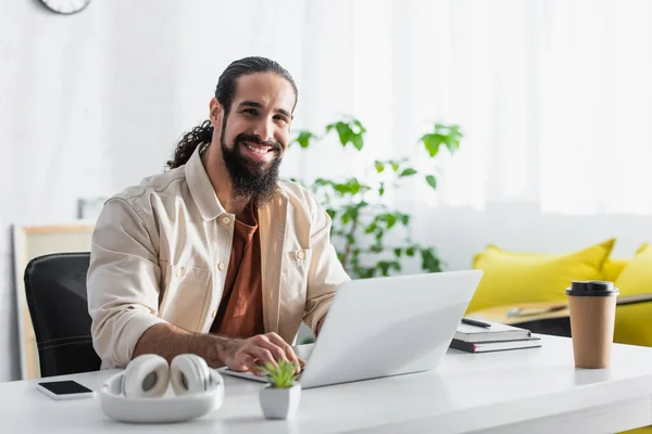 Heureux freelance latine regardant la caméra tout en travaillant sur ordinateur portable à la maison — Photo de stock