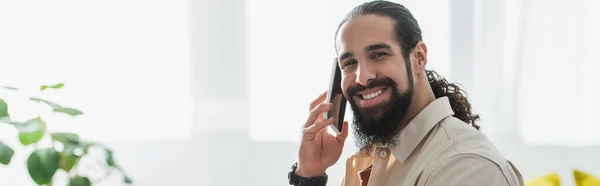 Barbudo latino hablando por teléfono móvil y sonriendo a la cámara en casa, pancarta - foto de stock