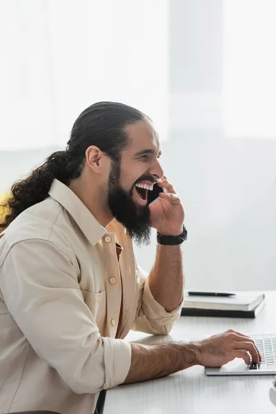 Emocionado freelancer hispano riendo mientras habla en el teléfono celular cerca del ordenador portátil - foto de stock