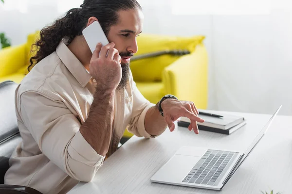 Bärtiger hispanischer Freiberufler zeigt auf Laptop, während er zu Hause auf dem Smartphone spricht — Stockfoto