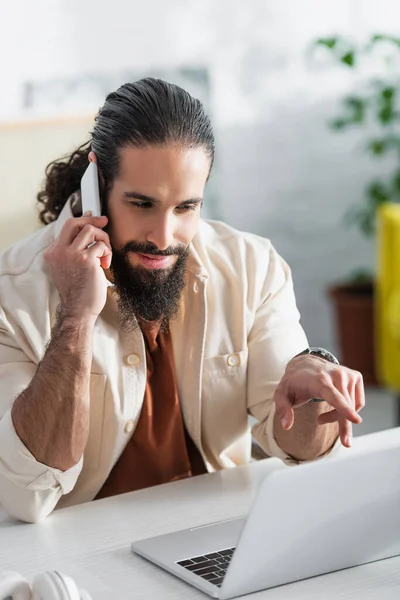 Freelancer hispano barbudo apuntando a una computadora portátil borrosa mientras habla por teléfono móvil - foto de stock