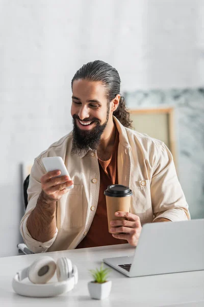 Alegre freelancer hispano sosteniendo café para llevar y mensajería en el teléfono inteligente en casa - foto de stock