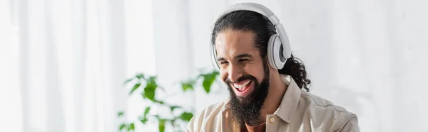 Hombre hispano emocionado riendo mientras escucha música en auriculares inalámbricos, pancarta - foto de stock
