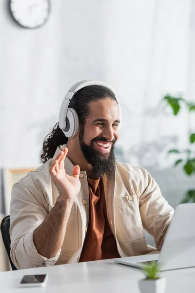 Freelancer hispânico feliz em fones de ouvido acenando a mão durante o bate-papo por vídeo no laptop borrado — Fotografia de Stock