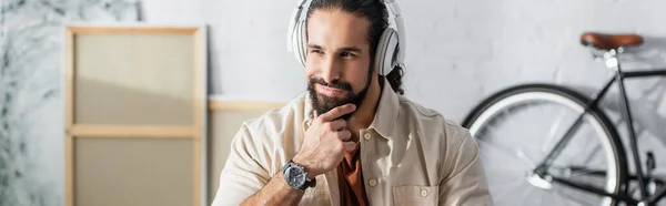 Sonriente y reflexivo hombre hispano tocando la barba mientras escucha música en auriculares, pancarta - foto de stock