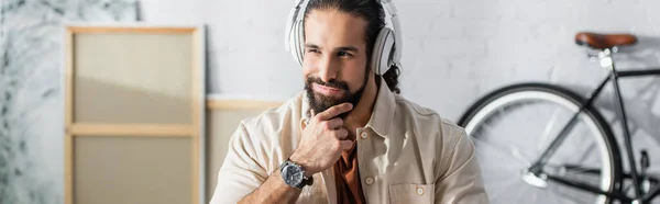 Lächelnder und nachdenklicher spanischer Mann, der den Bart berührt, während er Musik über Kopfhörer hört, Banner — Stockfoto