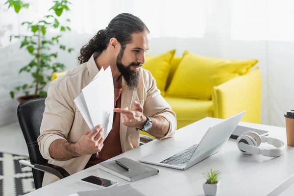 Bearded hispanic freelancer pointing at documents during video call on laptop at home — Stock Photo