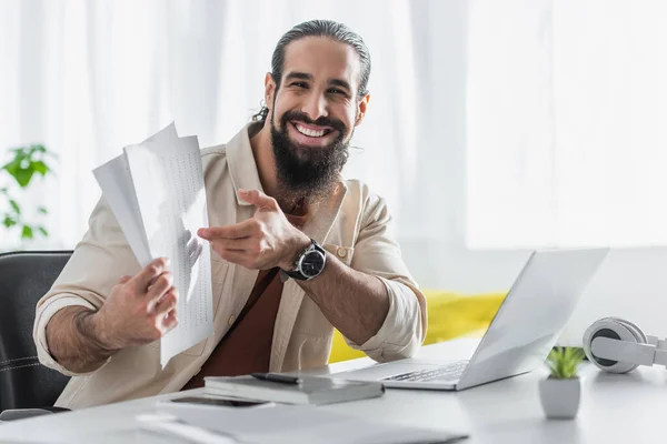 Freelancer latino positivo apuntando a los papeles mientras trabaja cerca de la computadora portátil en casa - foto de stock