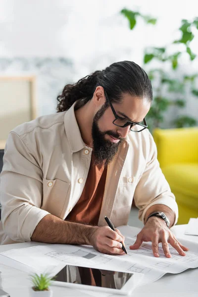 Arquitecto hispano barbudo en anteojos que escribe sobre plano mientras trabaja en casa - foto de stock