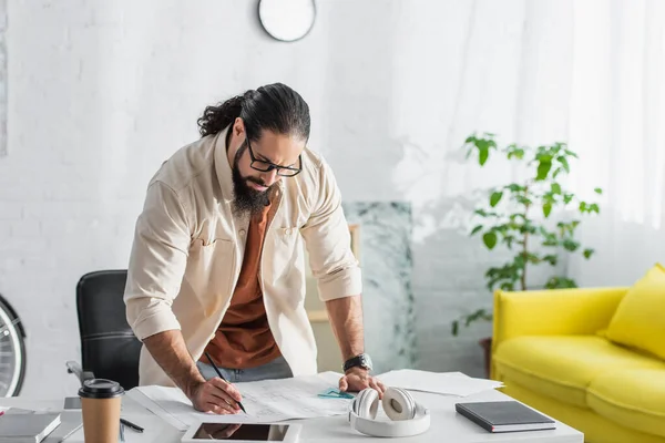 Bärtiger hispanischer Architekt schreibt auf Blaupause neben digitalem Tablet und Kopfhörer auf Schreibtisch — Stockfoto