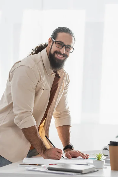 Heureux concepteur latin souriant à la caméra tout en tenant crayon près du plan à la maison — Photo de stock