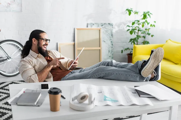 Designer ispanico che ride indicando tablet digitale mentre seduto con le gambe sulla scrivania a casa — Foto stock