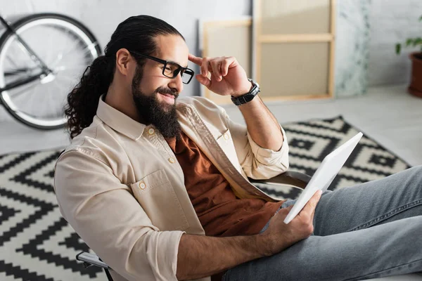 Spanischer Mann lächelt und berührt Kopf, während er digitales Tablet zu Hause benutzt — Stockfoto