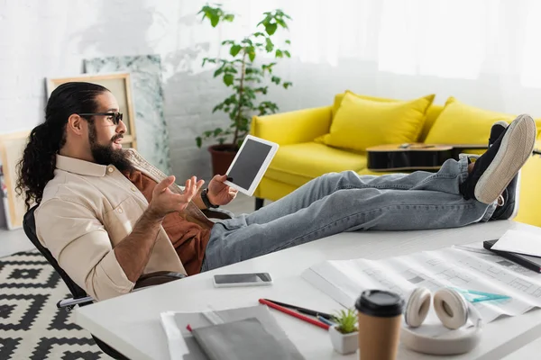 Designer latino surpreso sentado com as pernas na mesa de trabalho e gestos perto de tablet digital — Fotografia de Stock
