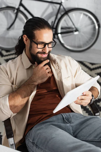 Hombre hispano con anteojos sonriendo y tocando la barba mientras mira la cámara - foto de stock