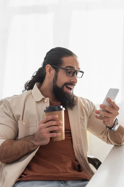 Amazed hispanic man with coffee to go looking at smartphone at home — Stock Photo