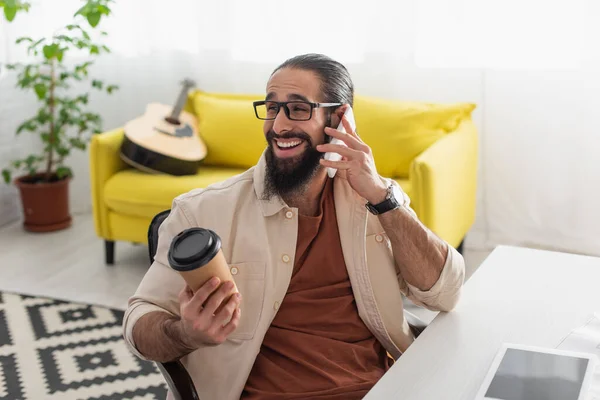 Hombre hispano feliz con café para ir a hablar en el teléfono inteligente cerca de sofá amarillo y la guitarra sobre fondo borroso - foto de stock