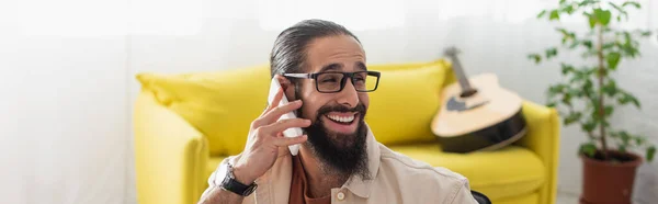 Hombre hispano feliz hablando en el teléfono inteligente cerca del sofá y la guitarra acústica sobre fondo borroso, bandera - foto de stock