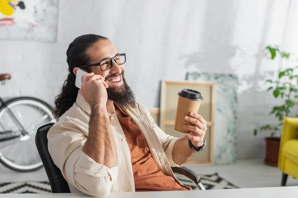 Alegre hombre hispano con bebida para llevar hablando por teléfono móvil en casa - foto de stock