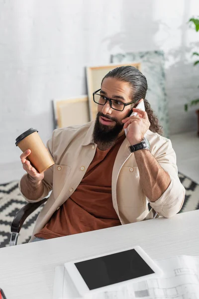 Surpris homme hispanique avec du café pour aller parler sur un téléphone portable près de tablette numérique avec écran blanc — Photo de stock