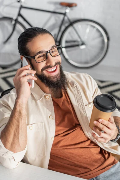 Homme latin joyeux avec du café pour aller parler sur smartphone près de vélo sur fond flou à la maison — Photo de stock