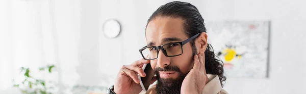 Thoughtful latin man in eyeglasses looking at camera while talking on mobile phone, banner — Stock Photo