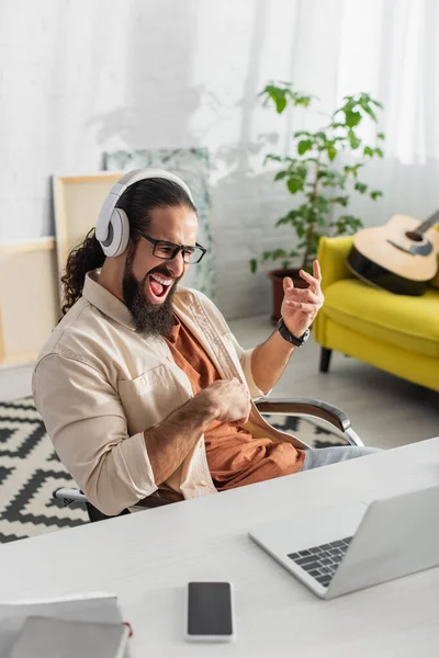 Emocionado freelancer hispano en auriculares gritando mientras escucha música cerca de la computadora portátil - foto de stock