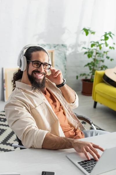 Freelancer hispano barbudo en auriculares sonriendo a la cámara mientras escucha música cerca de una computadora portátil borrosa - foto de stock