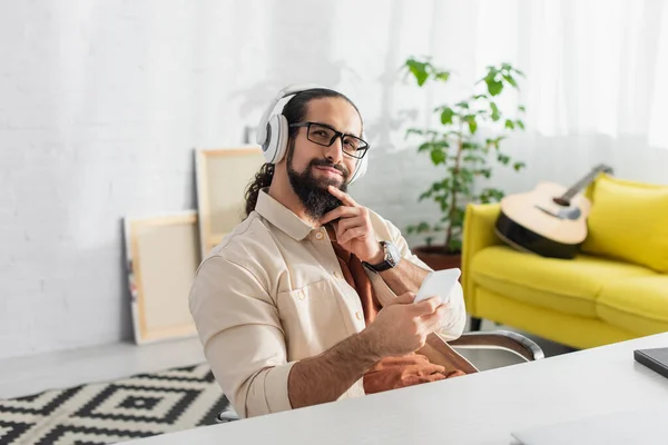 Hombre hispano soñador y sonriente con smartphone escuchando música en auriculares en casa - foto de stock