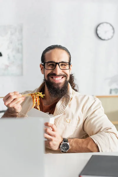 Freelancer hispânico alegre comer macarrão chinês perto de laptop borrado em casa — Fotografia de Stock