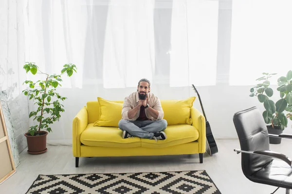 Ganzkörperansicht des hispanischen Mannes, der mit überkreuzten Beinen auf einer gelben Couch im modernen Wohnzimmer sitzt — Stockfoto