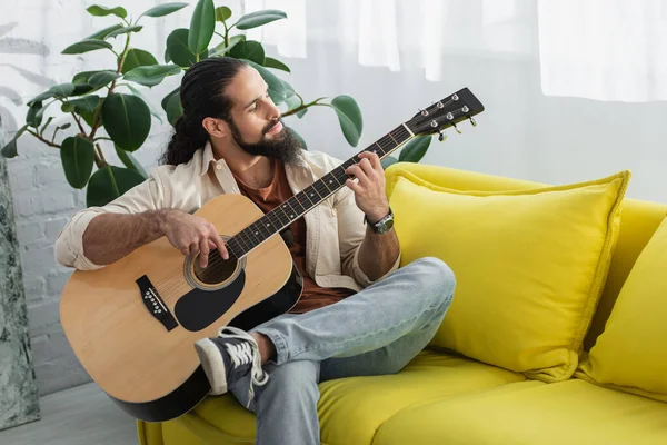 Hombre latino tocando la guitarra mientras está sentado en el sofá amarillo en casa - foto de stock