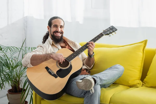 Felice uomo latino guardando la fotocamera mentre suona la chitarra sul divano giallo a casa — Foto stock