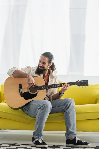 Hombre latino positivo sentado en la sala de estar en el sofá y tocando la guitarra - foto de stock